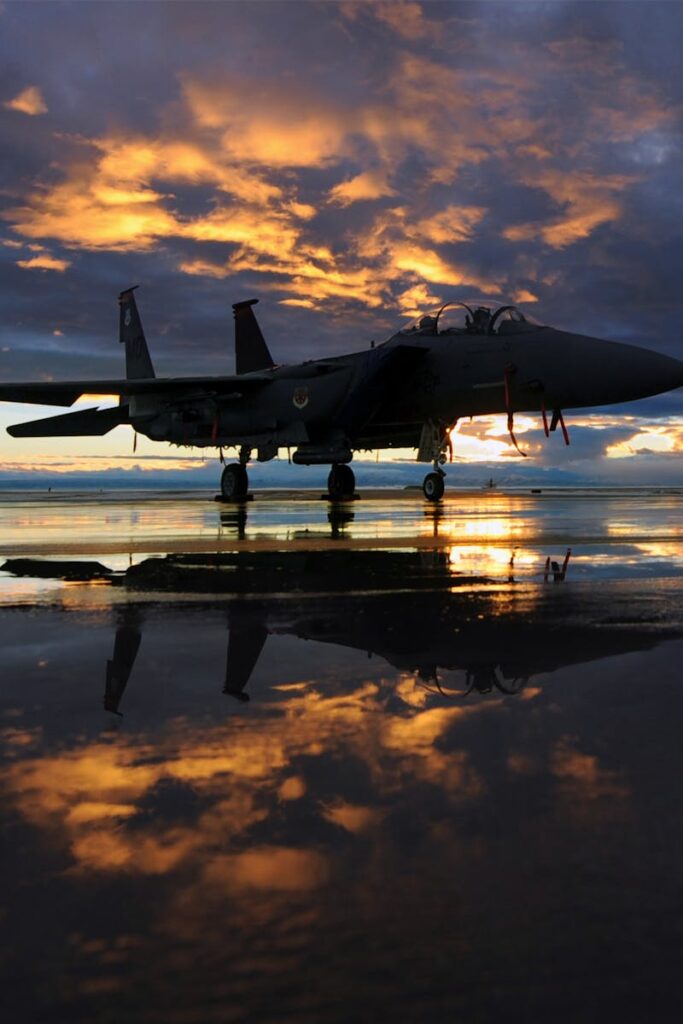 A military jet with striking sunset reflection on a wet tarmac, showcasing vibrant skies.
